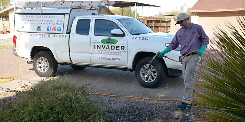 A pest control technician spraying a treatment from a hose beside a service pickup truck parked in a residential driveway.