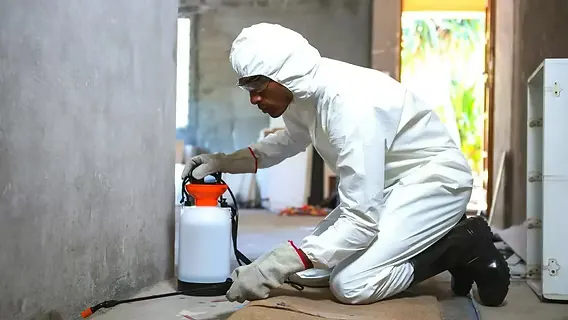 A pest control technician in white protective coveralls and a respirator kneeling with a sprayer inside a bare, unfinished structure.