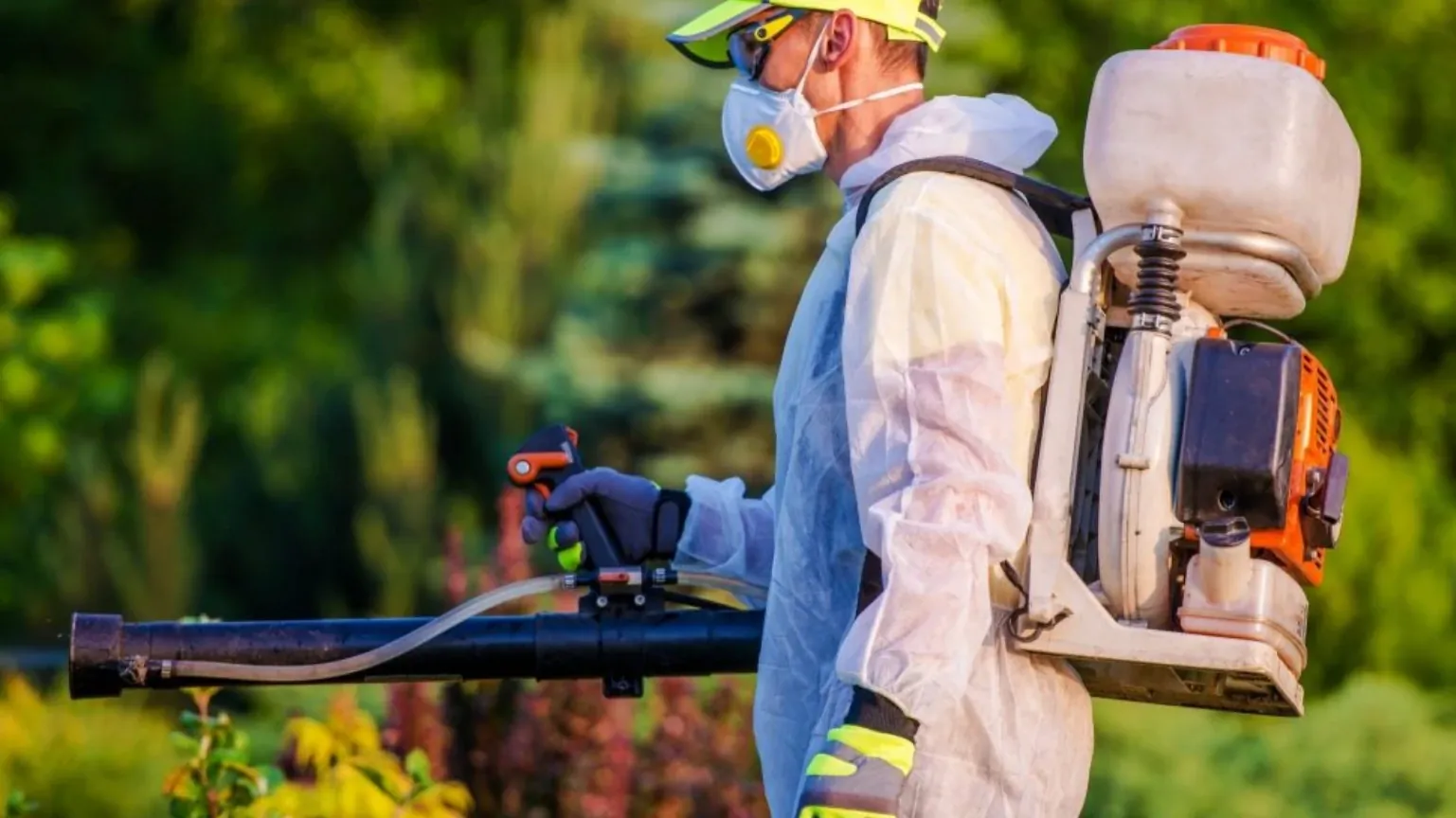 A pest control technician in white protective coveralls, a respirator, and gloves carrying a backpack mist blower while treating landscaping outdoors.