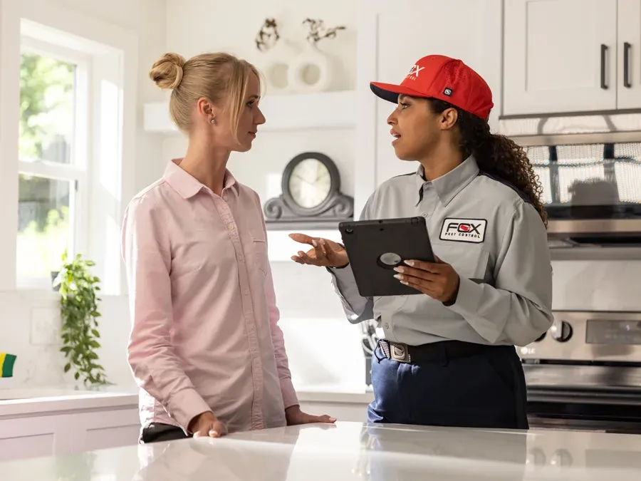 A pest control technician in a uniform holding a tablet while speaking with a customer in a kitchen.