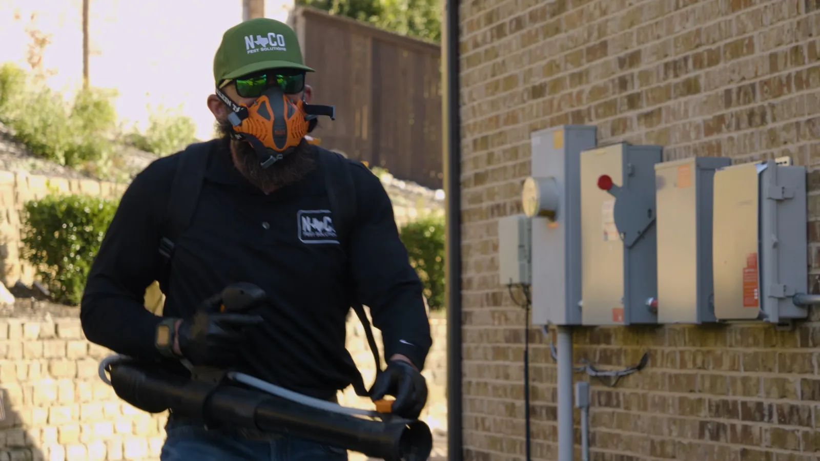 A pest control technician in a respirator and gloves carrying fogging equipment beside the brick exterior wall and utility meters of a building.